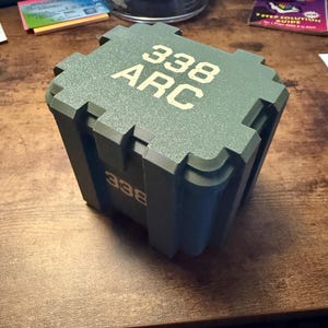 May include: A green box with the text "338 ARC" in a light tan color. The box has a textured surface and appears to be made of plastic. The box is sitting on a wooden surface.