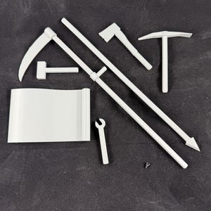 May include: A collection of white miniature tools, including a scythe, hammer, pickaxe, and wrench, arranged on a dark surface. The tools are likely for a miniature project or model.
