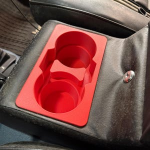 Könnte beinhalten: Ein leuchtend roter Getränkehalter-Einsatz mit zwei kreisförmigen Öffnungen, eingebettet in eine schwarze Leder-Autokonsole. Der Getränkehalter ist so konzipiert, dass er fest in die Konsole passt und einen sicheren Platz für Getränke bietet.