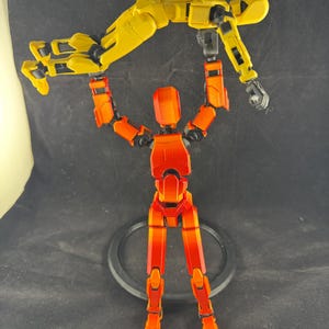 以下が含まれることがあります： 2つのパーツで構成された、可動式のアクションフィギュアセット。1つのフィギュアは黄色で、赤とオレンジのフィギュアによって持ち上げられています。フィギュアはダイナミックでアクション指向のポーズをとっています。フィギュアは黒い表面にあります。