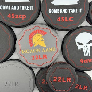 May include: A selection of black and grey circular items with different designs and text. Some have the words "COME AND TAKE IT" with "45acp" and "45LC" text. Others show a Spartan helmet, the text "ΜΟΛΩΝ ΛΑΒΕ" and "22LR".