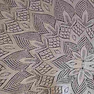 Op de afbeelding: Een crèmekleurig, rond kanten tafelkleed met een ingewikkeld bloemmotief. Het delicate opengewerkte patroon heeft herhalende blad- en geometrische vormen, waardoor een gedetailleerd en elegant textiel ontstaat. De randen zijn geschulpt, wat een decoratieve afwerking geeft.