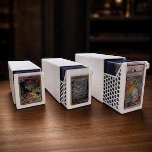 May include: Three white rectangular storage boxes for trading cards, each featuring a front-facing display slot with a honeycomb pattern on the sides. The boxes are arranged in a row, holding various collectible cards in protective plastic cases.