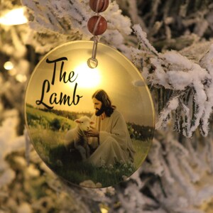 Może przedstawiać: Okrągła ozdoba choinkowa z napisem "The Lamb". Ozdoba przedstawia wizerunek Jezusa z barankiem na polu. Wisi na sznurku koralik&oacute;w na tle ośnieżonej choinki.