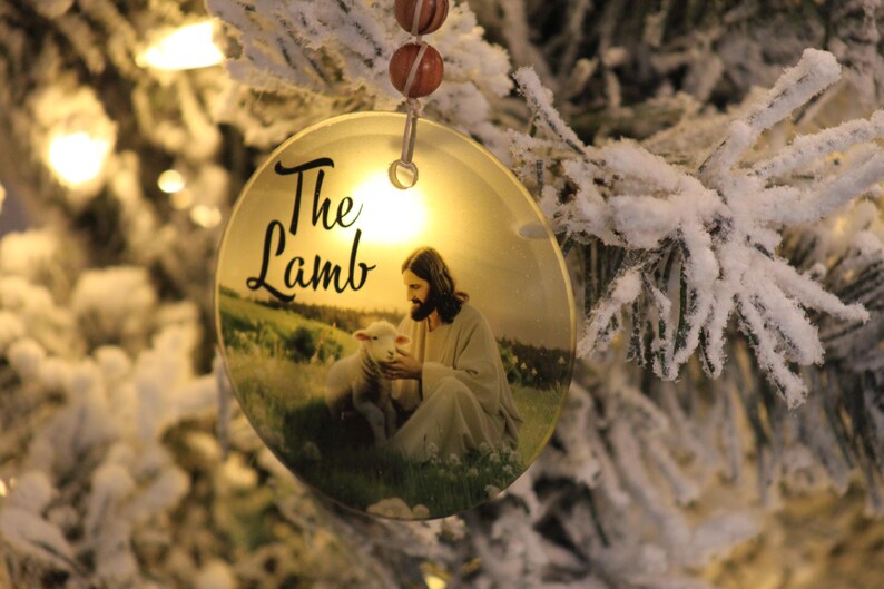 Może przedstawiać: Okrągła ozdoba choinkowa z napisem "The Lamb". Ozdoba przedstawia wizerunek Jezusa z barankiem na polu. Wisi na sznurku koralik&oacute;w na tle ośnieżonej choinki.