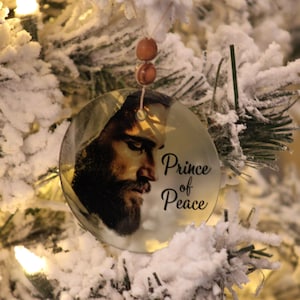 Może przedstawiać: Okrągła ozdoba choinkowa z wizerunkiem brodatego mężczyzny i napisem "Prince of Peace". Ozdoba zawieszona jest na sznurku z drewnianych koralik&oacute;w, na tle ośnieżonej choinki.