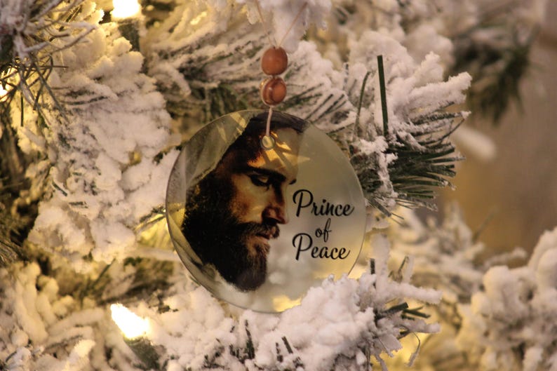 Może przedstawiać: Okrągła ozdoba choinkowa z wizerunkiem brodatego mężczyzny i napisem "Prince of Peace". Ozdoba zawieszona jest na sznurku z drewnianych koralik&oacute;w, na tle ośnieżonej choinki.