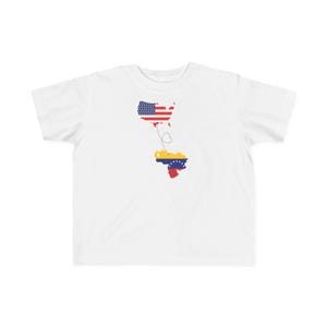 May include: White t-shirt featuring a map of the United States and Venezuela, with the flags of each country incorporated into the map outlines. A dotted line connects the two maps, symbolizing travel.