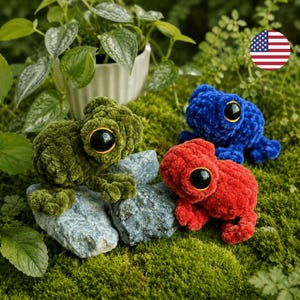Puede incluir: Tres juguetes de peluche con forma de rana en verde, rojo y azul, cada uno con grandes ojos negros y sentados sobre rocas grises. Las ranas están sobre un lecho de musgo verde, con una planta en maceta al fondo.