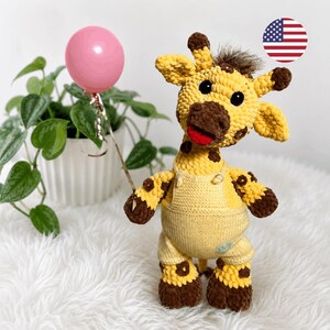 May include: A crocheted giraffe toy with yellow and brown fur, wearing light yellow knit overalls. The giraffe has black eyes, a red mouth, and a tuft of brown hair on its head. It holds a thin silver wire attached to a small pink balloon.