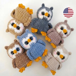 May include: A collection of handmade crochet owls in various colors, including brown, blue, and gray. Each owl has large, expressive eyes, a white face, and orange feet. The owls are arranged on a white surface.