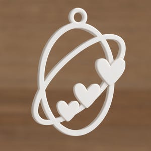 Puede incluir: Un colgante blanco con un diseño de forma orbital con tres corazones. El colgante tiene un lazo en la parte superior para colgarlo. El diseño es simple y elegante, adecuado para joyería o fines decorativos.
