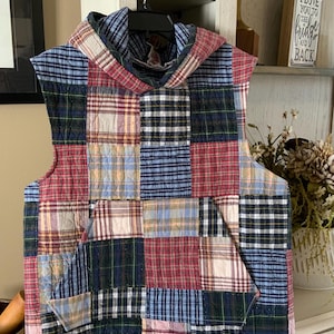 May include: A patchwork vest with a hood, featuring a variety of plaid patterns in red, blue, green, and white. The vest has a kangaroo pocket and is hanging on a black hanger.
