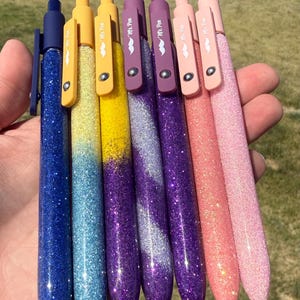 Puede incluir: Colección de siete bolígrafos con purpurina en una variedad de colores, incluyendo azul, amarillo, morado y rosa. Cada bolígrafo tiene un clip a juego y el texto "Mr. Pen". Los bolígrafos se sostienen en una mano sobre un fondo verde borroso.