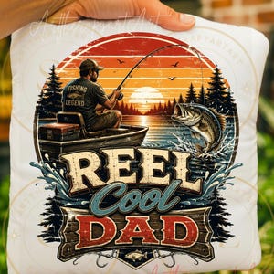 May include: White pillow with a fishing-themed graphic. The design features a man fishing from a boat at sunset, a leaping fish, and the text "REEL Cool DAD." The design includes trees, water, and a fishing rod.