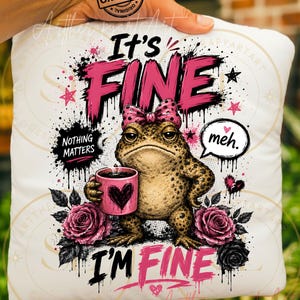 May include: A white square cushion featuring a graphic of a brown toad wearing a pink polka-dot bow, holding a pink mug with a heart. The text reads It's FINE, NOTHING MATTERS, meh., and I'M FINE. The design includes pink and black roses and stars.