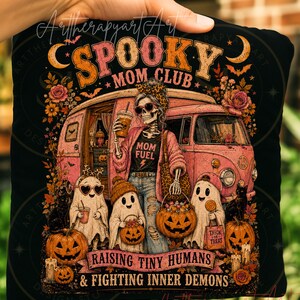 Può includere: Una t-shirt nera con la grafica di uno scheletro che indossa un cardigan rosa e occhiali da sole davanti a un furgone rosa con tre fantasmi. Il testo recita SPOOKY MOM CLUB, MOM FUEL, RAISING TINY HUMANS e FIGHTING INNER DEMONS.
