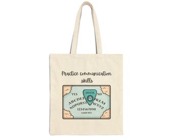 May include: Beige tote bag with a Ouija board design. The board is light blue with a planchette and the words "YES", "NO", and the alphabet. The text "Practice communication skills" is above the board.
