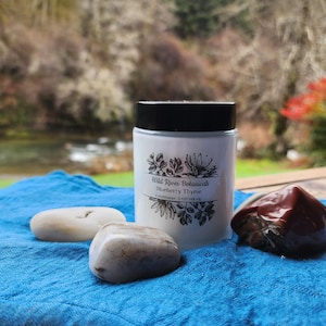 Puede incluir: Un tarro blanco con tapa negra, etiquetado "Wild River Botanicals Blueberry Thyme Conditioner 148 ml", está sobre una tela azul. Tres piedras lisas y una piedra marrón rojiza están dispuestas alrededor del tarro. El fondo muestra un río y árboles.
