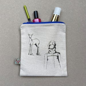 May include: A cream-colored canvas cosmetic bag with a blue zipper, featuring black line drawings of a deer and a person in a coat. The bag contains a green eyeliner pencil, a pink nail polish bottle, and a gold tube. A small patterned tag is attached.