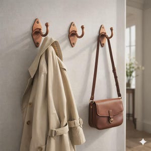 Könnte beinhalten: Drei kupferfarbene Wandhaken mit einem beigefarbenen Trenchcoat und einer braunen Leder-Umhängetasche. Die Haken sind an einer hellgrauen Wand befestigt und bieten eine praktische und stilvolle Aufbewahrungslösung.