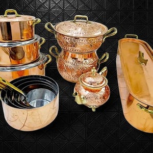 Puede incluir: Una colección de utensilios de cocina de cobre, que incluye ollas, sartenes y una tetera. Las ollas y sartenes tienen asas y tapas de latón. Los utensilios de cocina están colocados sobre una superficie negra.