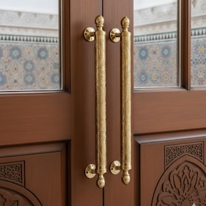 May include: Close-up of a wooden door with two ornate, gold-colored door handles. The handles are vertical, cylindrical, and feature intricate engraved patterns. The door is a rich brown color, with decorative carvings.
