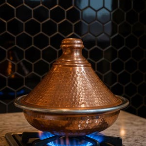 Può includere: Un tajine di rame con una trama martellata è posto su un fornello a gas con fiamme blu. La pentola ha un coperchio conico e una base larga, con un bordo argentato. Lo sfondo presenta piastrelle esagonali nere.