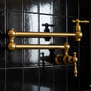 Puede incluir: Un grifo de llenado de ollas dorado, montado en la pared, con un brazo de doble articulación. El grifo tiene un diseño clásico con manijas decorativas y está montado en una pared de azulejos negros. El grifo es de latón.