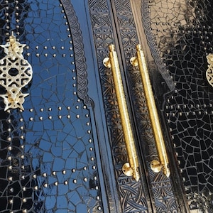 Op de afbeelding: Close-up van een zwarte deur met ingewikkelde gravures en goudkleurige hardware. De deur heeft twee lange, cilindrische handgrepen en een decoratieve grendel. Het oppervlak van de deur is bedekt met kleine, donkerblauwe mozaïektegels en bezet met kleine, goudkleurige klinknagels.