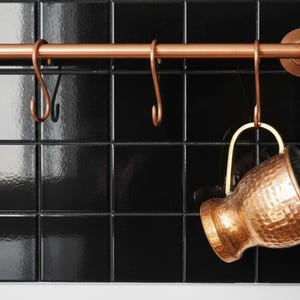 Puede incluir: Barra de cocina de color cobre con ganchos, montada en una pared de azulejos negros. Una taza de metal martillado de color cobre cuelga de uno de los ganchos. La barra y los ganchos tienen un acabado brillante.