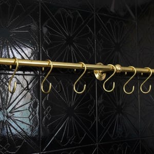 May include: A gold-coloured kitchen rail with eight S-shaped hooks, mounted on a black tiled wall. The rail has decorative end caps and mounting plates. The hooks are evenly spaced along the rail, designed for hanging kitchen utensils or other items.