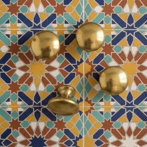 May include: Four brass cabinet knobs and pulls against a colorful tile background. The knobs are round and the pull is oval, all with a polished finish. The tile features geometric patterns in blue, yellow, brown, and teal.