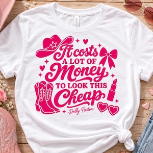 May include: A white short-sleeved t-shirt featuring a bright pink graphic print that reads It costs A LOT OF MONEY TO LOOK THIS Cheap -Dolly Parton. The design includes icons of a cowboy hat, a bow, cowboy boots, lipstick, and heart shapes.