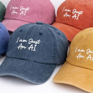 May include: Several baseball caps in shades of pink, orange, blue, and yellow. Each cap features the white embroidered text "I am Just An AI" across the front, a casual and modern design.