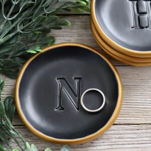 May include: A black ring dish with a gold rim and the letter "N" embossed on the surface. A silver ring is resting in the dish.
