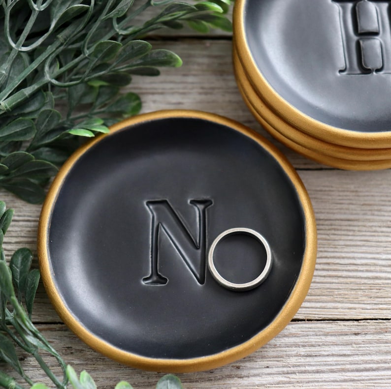 May include: A black ring dish with a gold rim and the letter "N" embossed on the surface. A silver ring is resting in the dish.