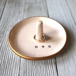 May include: A pink ceramic ring dish with a gold rim. The dish has a small, raised ring holder in the center and the words "a + e" written on the bottom.