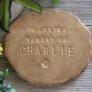 May include: A tan ceramic pet memorial plaque with an irregular shape. The embossed text reads "IN LOVING MEMORY OF CHARLIE", accompanied by a paw print. The plaque is presented on a wooden surface.