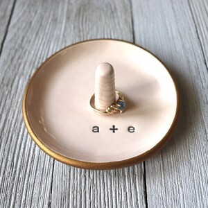 May include: A pink ceramic ring dish with a gold rim and a small white ceramic ring holder. The dish has the words "a + e" written on it.