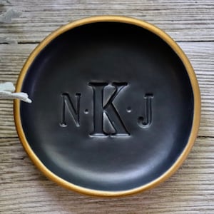 Puede incluir: Un plato para anillos negro con un borde dorado y las iniciales "N.K.J." grabadas en el centro.