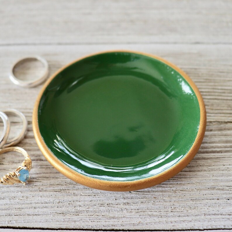 Op de afbeelding: Een klein, rond schoteltje met een groene binnenkant en een gouden rand. Het schoteltje staat op een houten oppervlak.