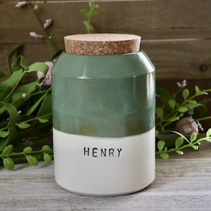 May include: A two-toned ceramic jar with a cork lid. The top half is a muted green, and the bottom half is white, with the name "HENRY" printed in black. The jar is cylindrical and sits on a wooden surface.