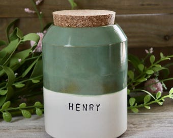 May include: A two-toned ceramic jar with a cork lid. The top half is a muted green, and the bottom half is white, with the name "HENRY" printed in black. The jar is cylindrical and sits on a wooden surface.