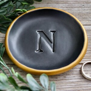 May include: A black ceramic ring dish with a gold rim and the letter "N" embossed in the center.