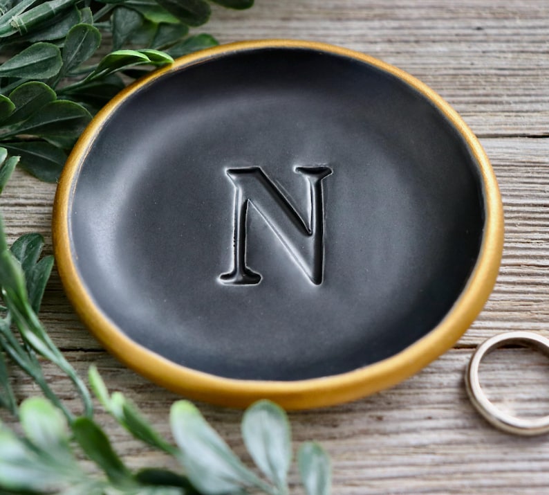 May include: A black ceramic ring dish with a gold rim and the letter "N" embossed in the center.