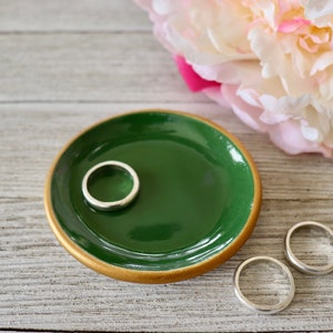 Op de afbeelding: Een groene keramische ringenschotel met een gouden rand. De schotel staat op een houten oppervlak en er liggen drie zilveren ringen op het oppervlak.