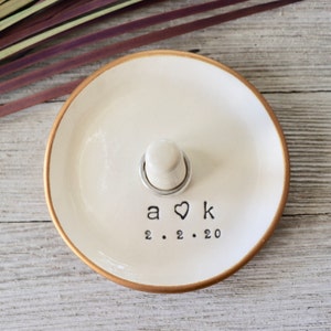 May include: White ceramic ring dish with a gold rim. The dish has a small, white ring holder in the center and the initials "A ♥ K" and the date "2.2.20" written on the bottom.