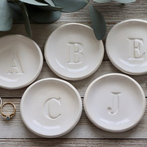 May include: Five white ceramic ring dishes with raised letters A, B, C, E, and J. The dishes are arranged on a light wood surface.