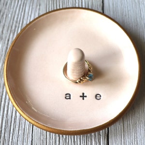 May include: A pink ceramic ring dish with a gold rim and a small white ceramic ring holder. The dish has the text "a + e" written on it.